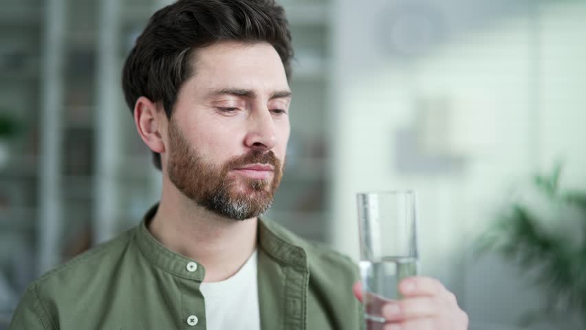 Happy handsome man drinks water from a glass with closed eyes sitting in living room at home. Satisfied smiling bearded male feeling relieved, enjoying clean cool drink, quenching his thirst. Close up