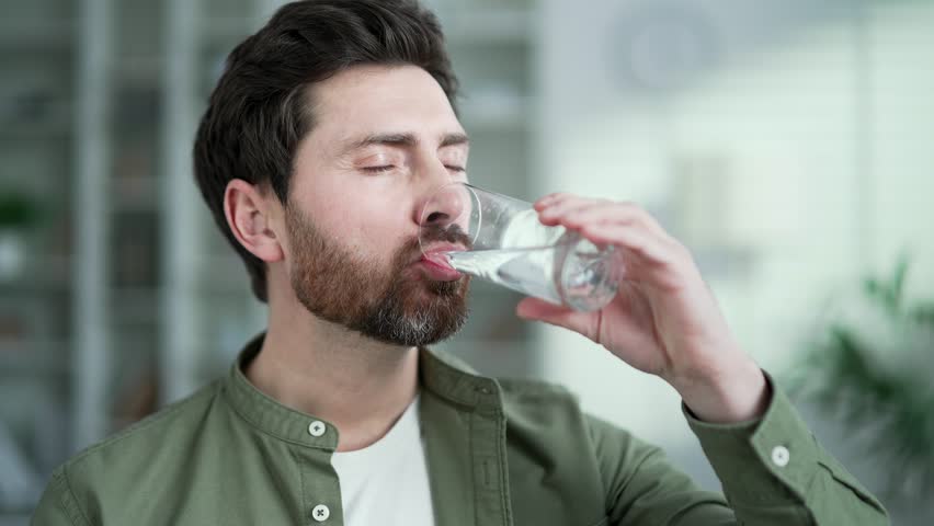 Happy handsome man drinks water from a glass with closed eyes sitting in living room at home. Satisfied smiling bearded male feeling relieved, enjoying clean cool drink, quenching his thirst. Close up