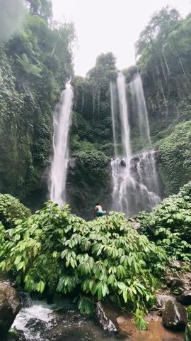 man stands on a rock overlooking a majestic waterfall surrounded by lush greenery at Sekumpul Waterfall, Bali, Indonesia. sstkVertical