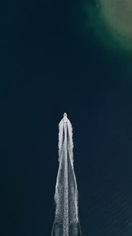 Aerial view of a lone boat speeding across the calm, dark blue ocean, leaving a trail of white waves behind. The simplicity and tranquility of the vast sea contrast with the boat