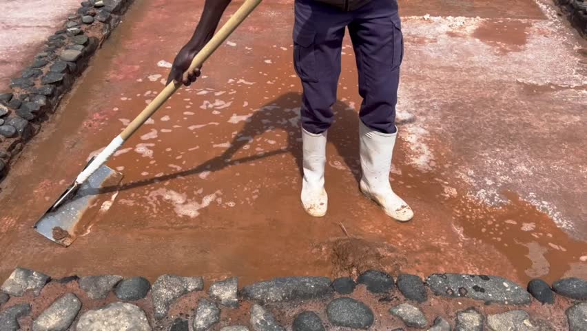 A man is cleaning a muddy area with a shovel and a broom. The area is filled with rocks and dirt, and the man is wearing boots. The scene has a somewhat gloomy and dirty atmosphere