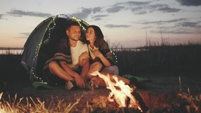 A couple is hugging next to their tent, both wrapped in a warm blanket and gazing at the campfire. Evening is turning into night, and string lights are hanging on the tent, creating a cozy atmosphere. - Powered by Shutterstock - Get 15% off with code: PIKWIZARD15