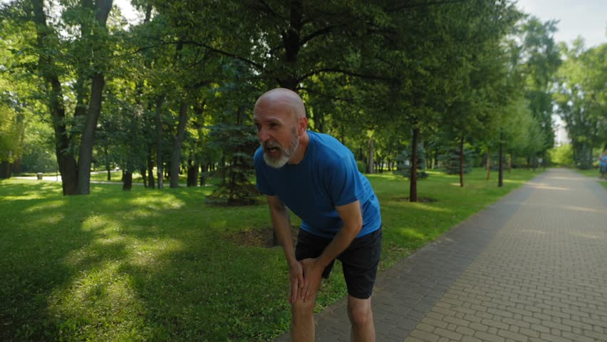 Active motivated athletic fit middle aged male runner suffering from knee injury, holding and massaging kneecap, trying to relieve pain while practicing outdoor jog workout.