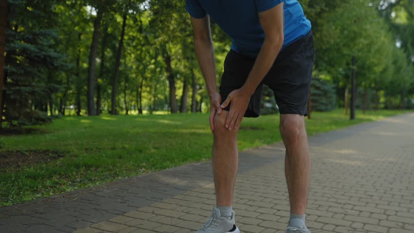 Low section of athletic fit middle aged man jogger massaging injured knee, trying to relieve physical ache while running in summer park.