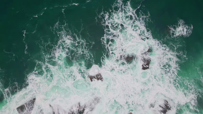 Stormy sea splashing at volcanic coastal stones top view. Dangerous ocean water crashing of rough coast rocks in super slow motion. Aerial shot scenic foamy waves hitting cliffside making white foam - Powered by Shutterstock - Get 15% off with code: PIKWIZARD15