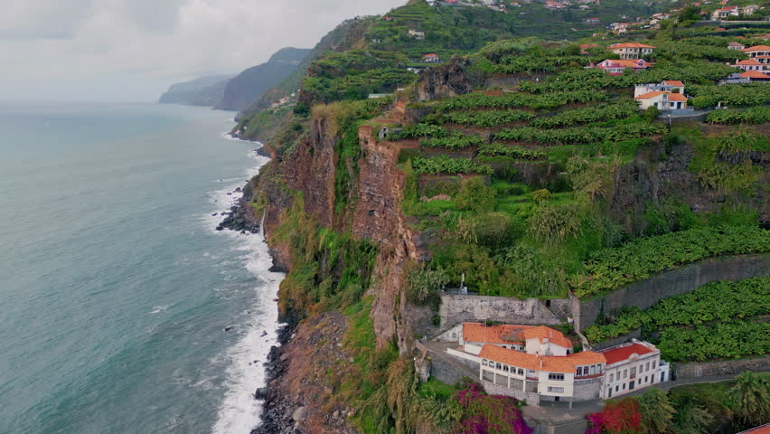 Aerial view coastal villa on green terraced cliffs. Beautiful houses on lush hills overlook picturesque ocean waves. Scenic foamy sea crashing rocky coast. Calm rural landscape perfect for vacation.