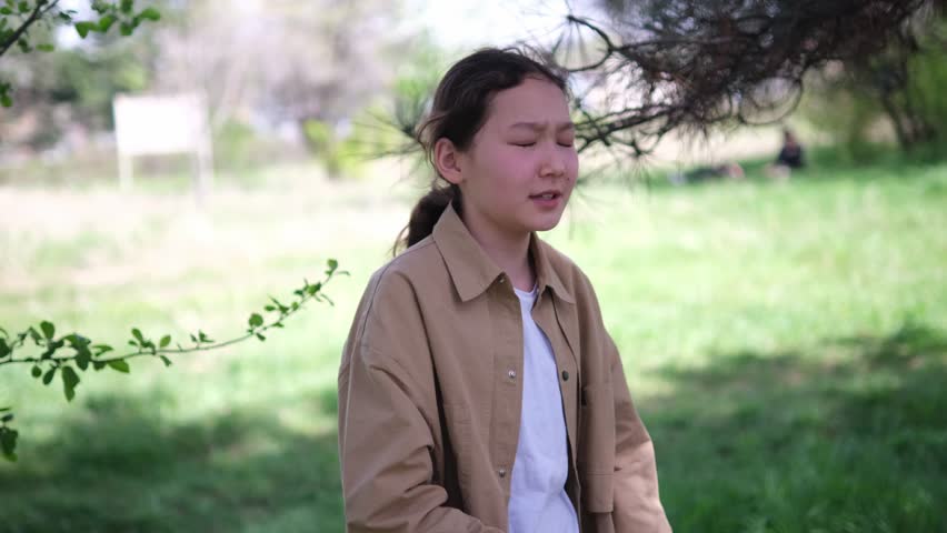 An Asian girl in a beige jacket and white shirt having fun outdoors. She is surrounded by green foliage and sunlight, creating a lively and cheerful scene. Outdoors activity, summer park