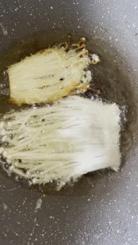 A close-up view of thinly sliced Enoki mushrooms partially submerged, frying in hot oil in a wok, with some pieces already turning golden brown.