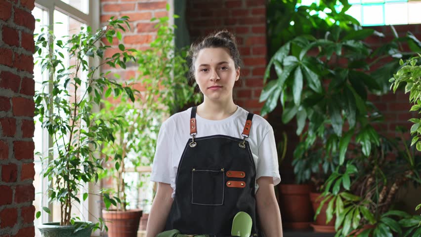 Running of own business. Young woman florist wearing apron in botanical store with green plants. Happy small business owner working at flower shop standing surrounded by plants. Small business