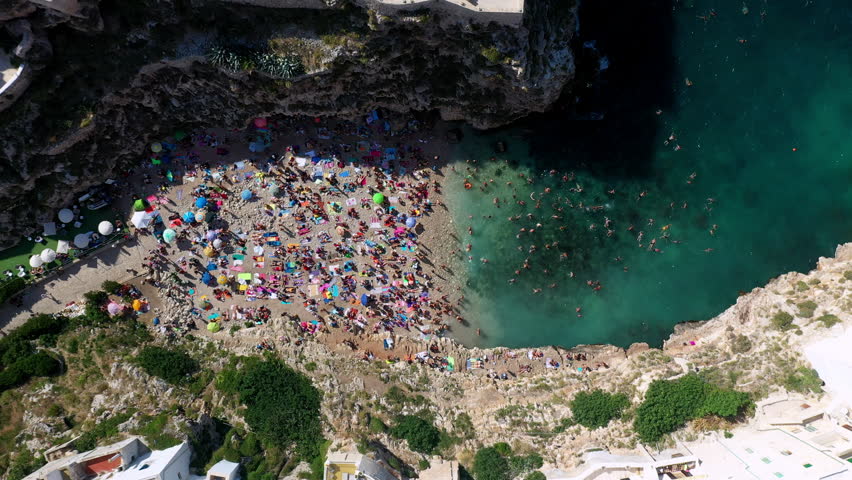 Bustling drone view of Lama Monachile beach on a sunny day in Puglia, Italy. People on the beach enjoying the clear blue waters and stunning cliffs. Rising aerial shot.