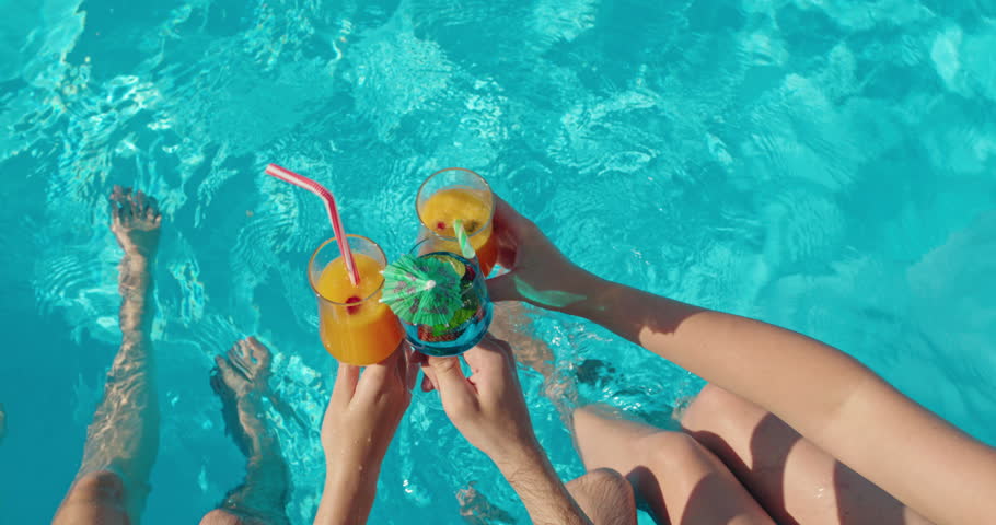 People clinking cocktail glasses against the water surface of a pool, top view