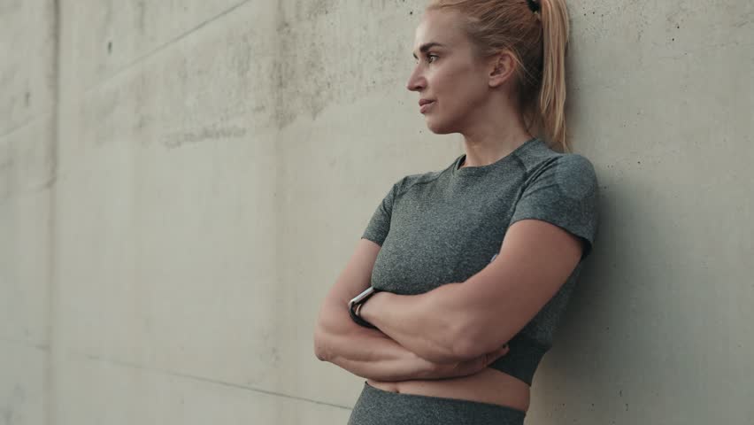A confident athletic woman rests against a concrete wall in her activewear, reflecting on her outdoor boxing training as the sunset approaches.