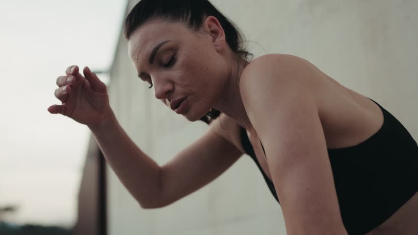 Exhausted athletic woman taking a break from running, leaning on a concrete wall outdoors with a focus on recovery.