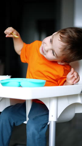 Lovey cute baby boy sits bent to the side in his high chair. Caucasian toddler having meals at home. Vertical video.