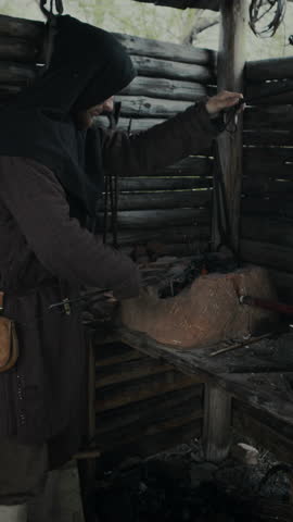 Medium vertical shot of male medieval village blacksmith in black hood heating piece of metal in forge and blowing on coals in fire with bellows