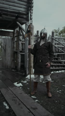 Full vertical portrait of young Caucasian soldier in metal helmet and brown leather tunic standing guard by gates of fortress, holding spear, looking around then at camera