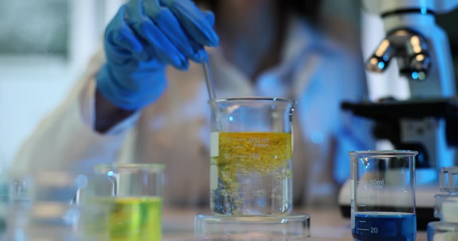 Lab worker stirs liquid with oil in beaker at workplace slow motion. Female scientist in latex gloves prepares ingredients for experiment