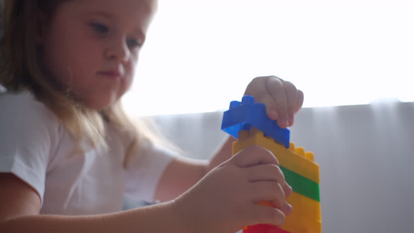 Selective focus of focused caucasian child play in constructor cubes at home. Cute little girl play colorful toys, blocks and cars on table in indoors kindergarten. Preschool education concept