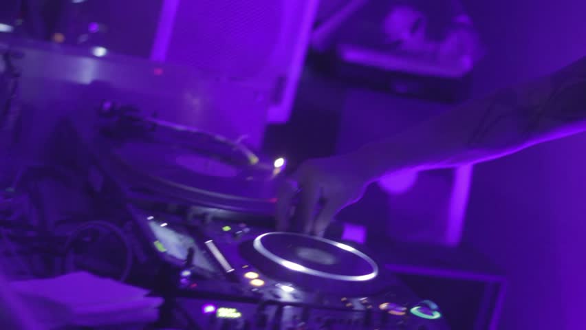 DJ tweaks turntable and mixer, controls music in nightclub.