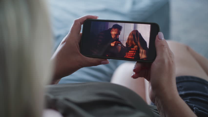 Adult woman watches action movie using mobile phone while lying on couch. Smartphone screen with displayed thriller film. Caucasian woman rests at home on the weekend. Lifestyle concept. Close Up.