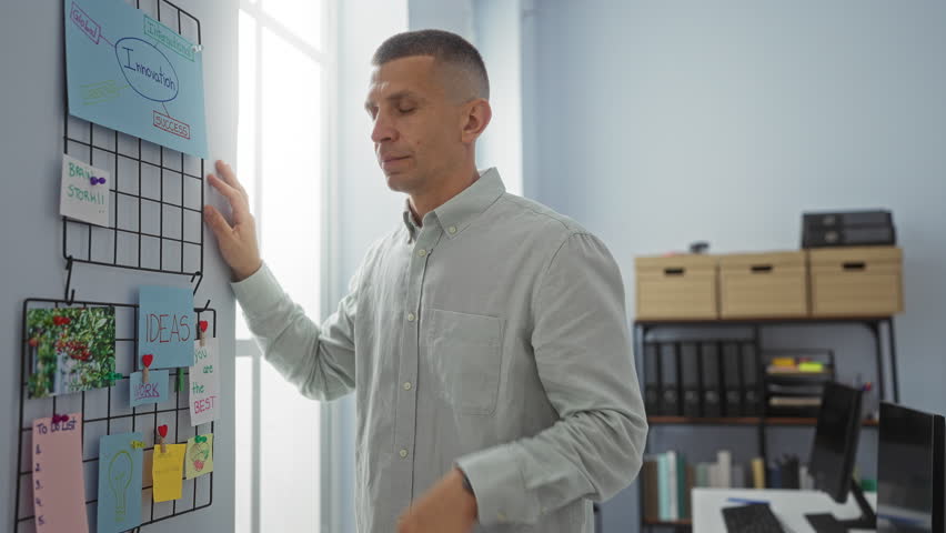 Young man in an office, looking stressed and frustrated, standing near a large window and a colorful idea board, with a modern interior featuring bookshelves and a computer system around