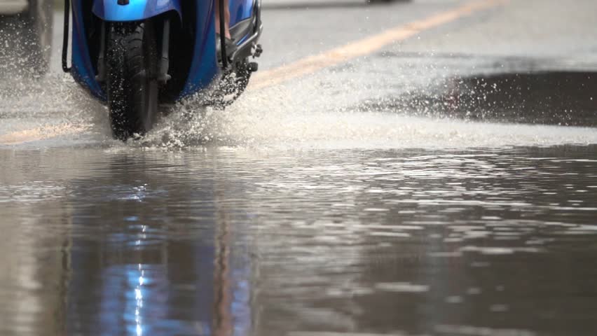 A motorcycle is driving through a flooded street. The water is deep and the motorcycle is splashing water everywhere. The scene is chaotic and dangerous, as the motorcycle could easily lose control