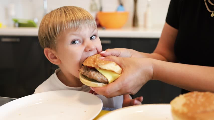 Happy mom and son kid eating in home kitchen together, having fun. little boy eating a hamburger. Child greedily with pleasure bites lifestyle a big burger in the kitchen at home. 