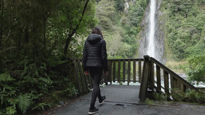 Young caucasian woman in a jacket walking and looking at a waterfall in a forest at Thunder Creek Falls, West Coast, New Zealand.
