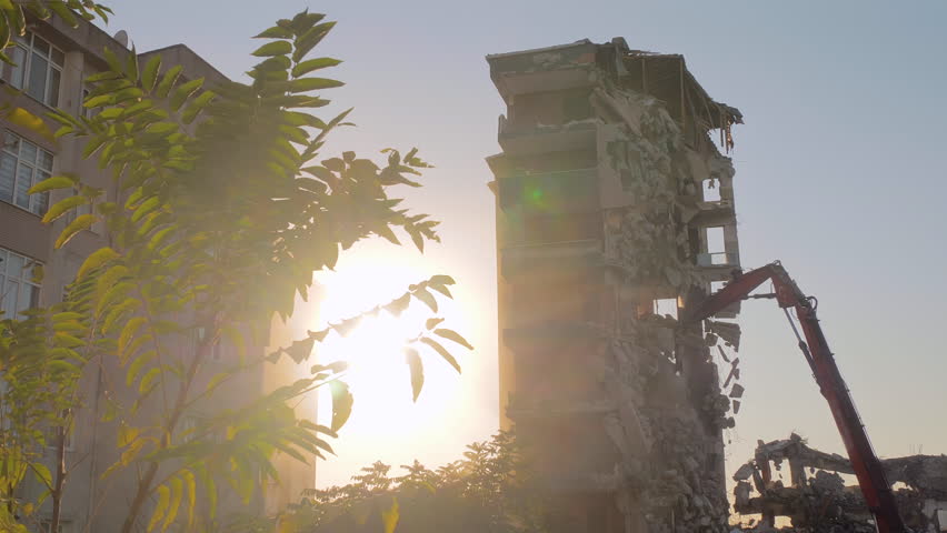 Excavator with hydraulic crusher is tearing down the old typical Turkish apartment, residential building in Istanbul, Turkey at sunset. Setting sun is shining through leaves: sun lens flares