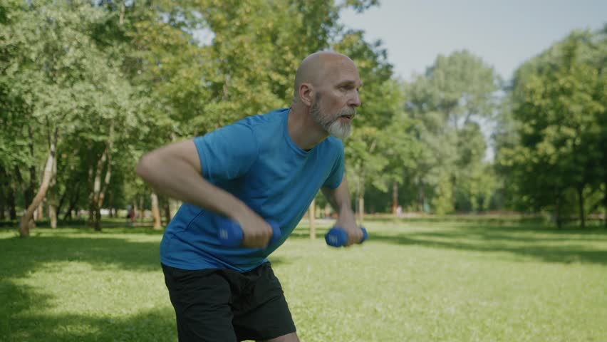 Portrait of determined motivated handsome sporty fit mature man in activewear practicing weight training , doing bent over dumbbell rows workout while exercising in public park.