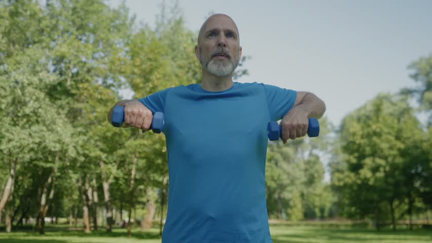 Determined sporty fit mature man exercising dumbbell upright row, strengthening deltoids, trapezius, and triceps while practicing outdoor sports training.