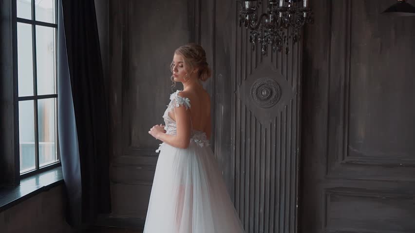 Luxurious young blonde woman stands at window waiting love, dark classic interior room. Bride girl in white wedding day dress, bare back open. Professional hairstyle, fantasy fairy princess style 