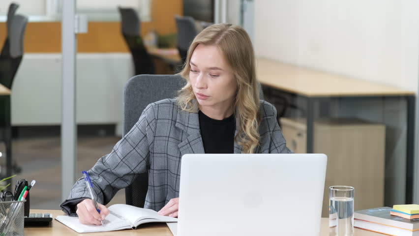 A professional employee is seen deeply engaged with his tasks, writing notes beside her laptop. Office worker busy on her desk, portrait concept. Planning.