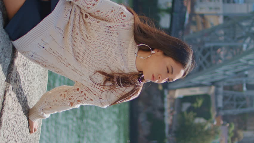 Lazy girl sitting river bank on sunny day closeup. Happy tourist lady enjoying spring vacation relaxing at stoned promenade vertical view. Pretty woman traveler admiring marine landscape on weekend.