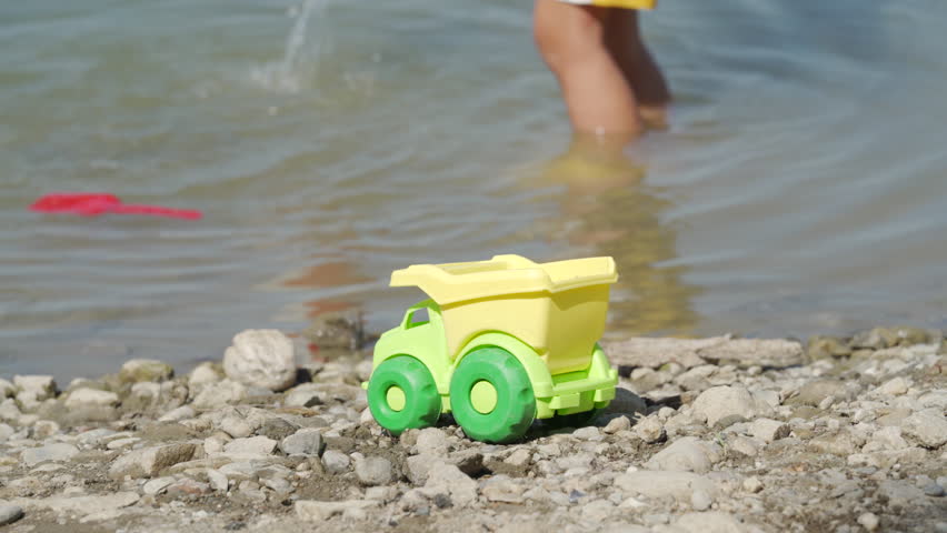 Small child playing with toys near water on lake shore in summer. Little boy wearing yellow sun hat in nature. Simferopol, Crimea, Russia 2024.