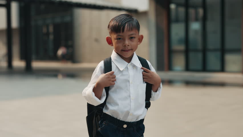 Small Asian boy dressed in white shirt stands in school courtyard looking at camera. Portrait of smiling schoolboy with large black backpack stands near main building of school waiting for classmates