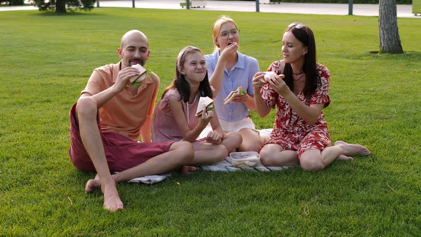 Portrait of a happy family with teenage children on the green grass in the park, they eat fresh sandwiches together. Family picnic in summer in a modern park.