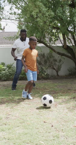 Vertical video: African American father and son are playing soccer. Both wearing casual clothes, father in white, the son in orange and blue