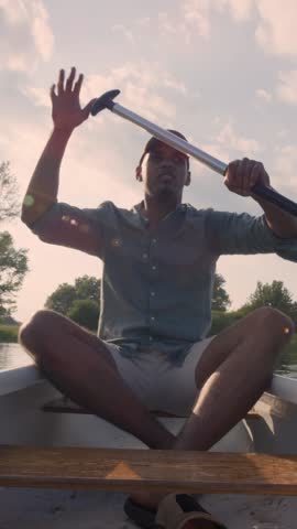 Young African American man canoeing, paddling, and moving canoe through the lake water, enjoying the natural environment, low angle vertical shot.