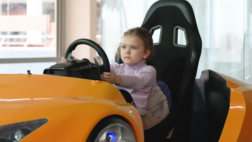 A little girl in an entertainment center with slot machines sits in a car game simulator and holds the steering wheel with her hands