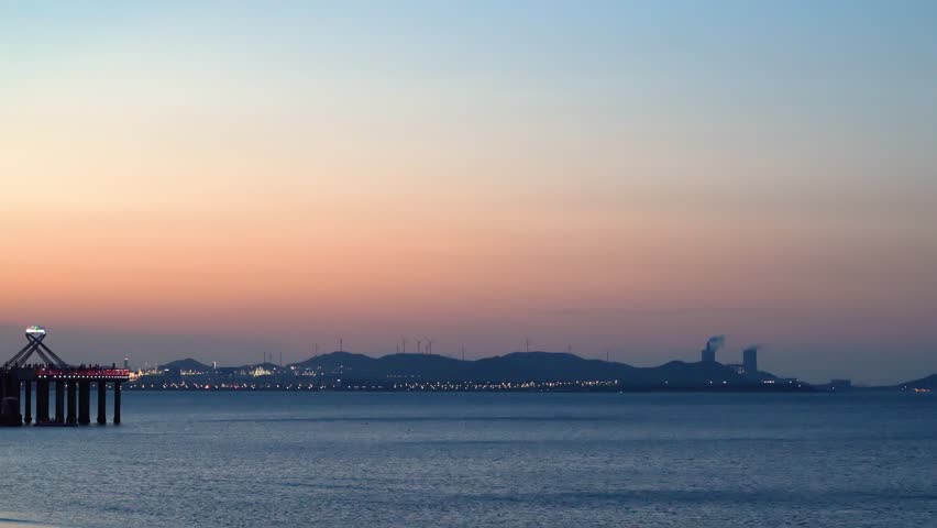 Wind turbines along the coast of a Chinese city at sunset and industrial exhaust chimneys, city lights, Jinshatan Beach Park, Chinese Yantai city