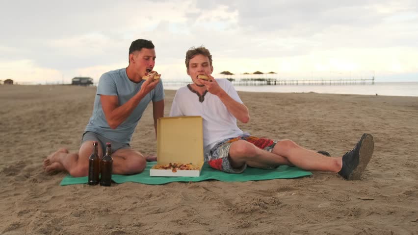 Gay couple having picnic eating pizza on beach during summer weekend, romantic relationship. Slow motion.