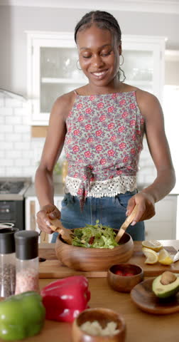 Vertical video: African American young woman preparing salad in kitchen. She has braided hair, wearing floral top, mixing greens in a wooden bowl