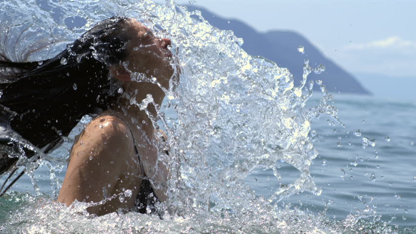 Woman tossing her head back in slow-motion in a lake, creating a stunning splash, vibrant and refreshing moment on a sunny day with mountains in the background