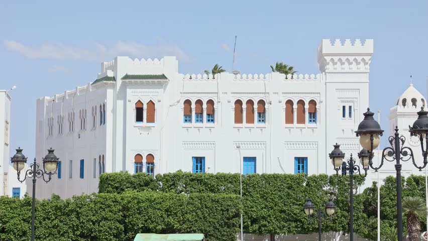 Sunlit elegant white building of Ministry of Religious Affairs in Tunis city surrounded by greenery at central Kasbah Square, with traditional Tunisian architectural elements and vivid blue shutters