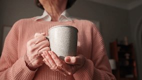 Camera view of elderly woman with stylish pink sweater holding cup of drink with both hands. Warming hands in cold season. Enjoying hot morning drink. Drinking coffee or tea. Harmony. - Powered by Shutterstock - Get 15% off with code: PIKWIZARD15