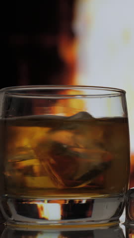 Glass and bottle of whiskey, scotch or bourbon stands on a table against background of a fireplace with a flame. Alcoholic drink and home comfort concept. vertical footage