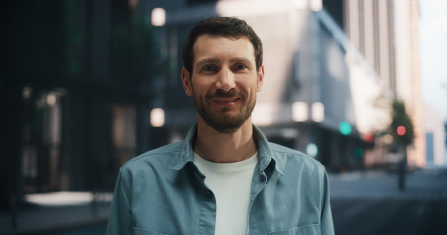 Handsome Caucasian Man Raises His Eyes and Smiles Warmly at the Camera on a Quiet City Street, Creating a Friendly and Inviting Urban Atmosphere.
