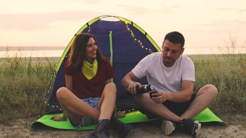 A couple is sitting next to their tent by the shore of a sea or lake. The guy is pouring tea or coffee from a thermos, and a string of lights is hanging on the tent. They are enjoying a peaceful