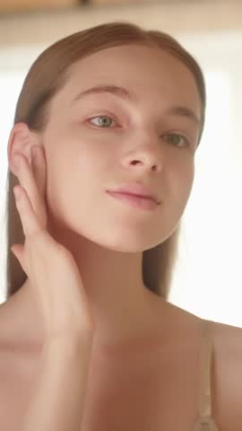 Vertical video. Young woman applying makeup foundation looking in the mirror. Young adult woman with blonde hair making her skin soft and clean with a natural skin care cosmetic Slow motion Beauty ads